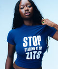 Studio portrait of a person wearing the Topicals navy Stop Staring At My Zits tee against a light blue background.