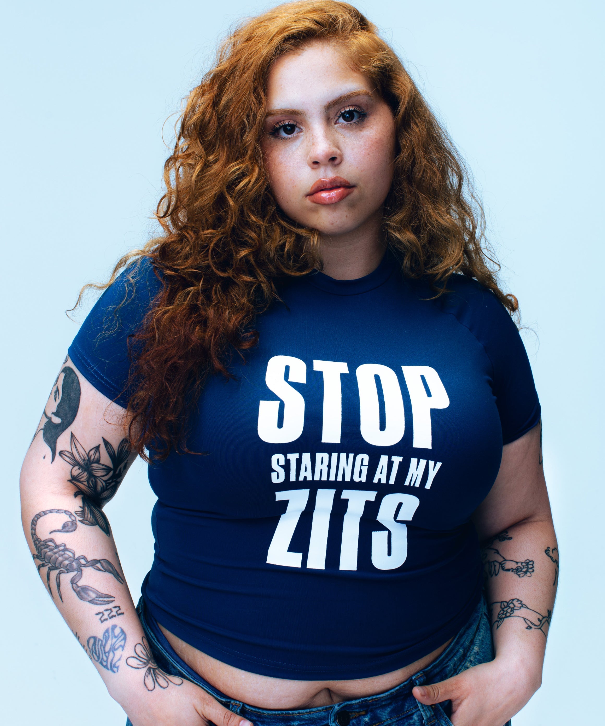 Studio portrait of a person with curly hair wearing the Topicals Stop Staring At My Zits tee against a blue backdrop.