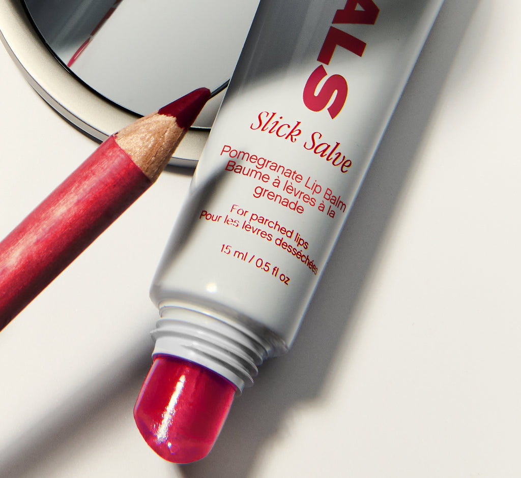 Moisturizing lip balm from Topicals Slick Salve in Pomegranate shown uncapped next to a red lip liner pencil and a round mirror on a white surface.