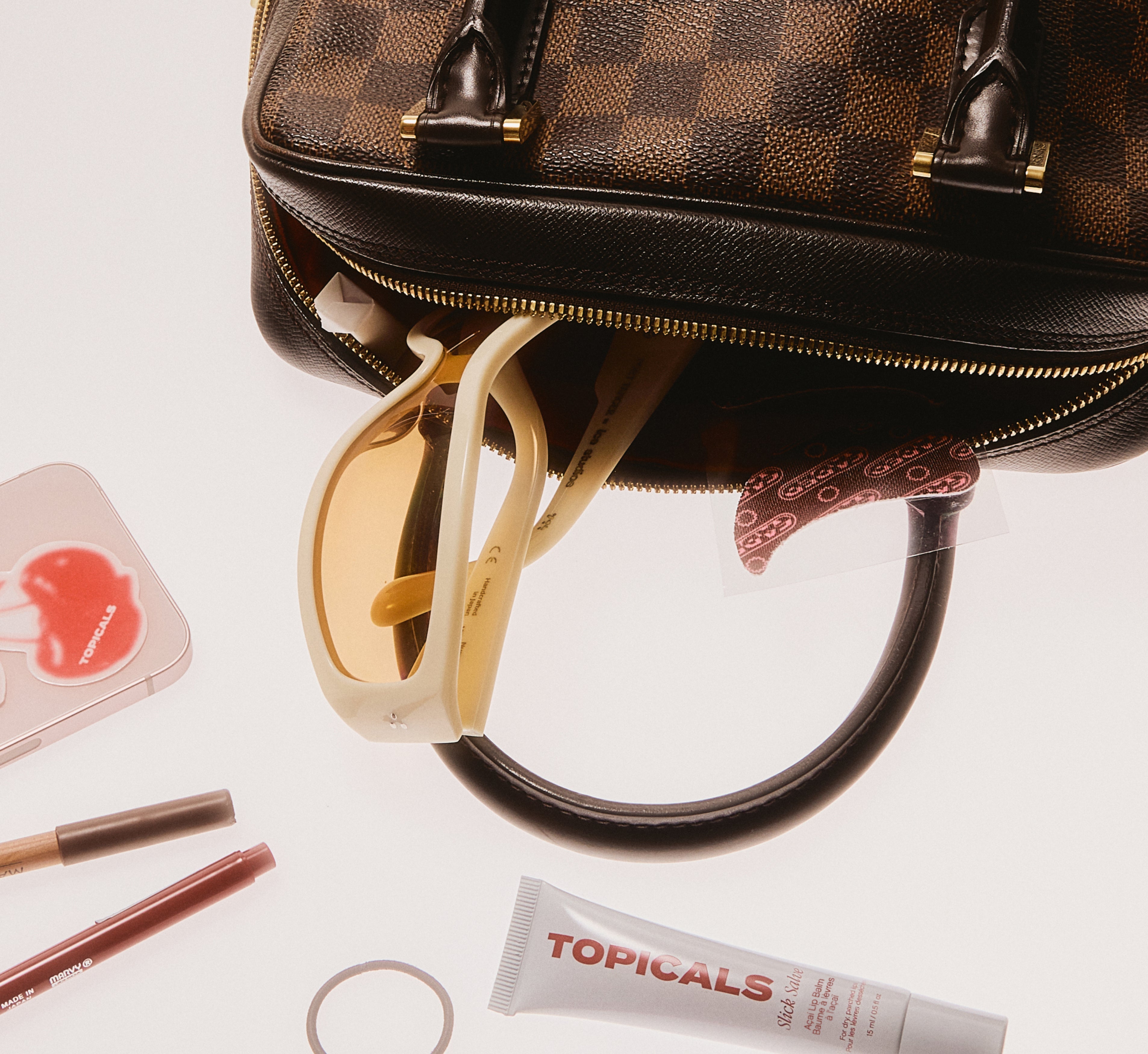 Brightening under eye patches from Topicals Faded peeking out of a designer handbag surrounded by sunglasses, lip liners, and a Topicals Slick Salve lip balm tube.