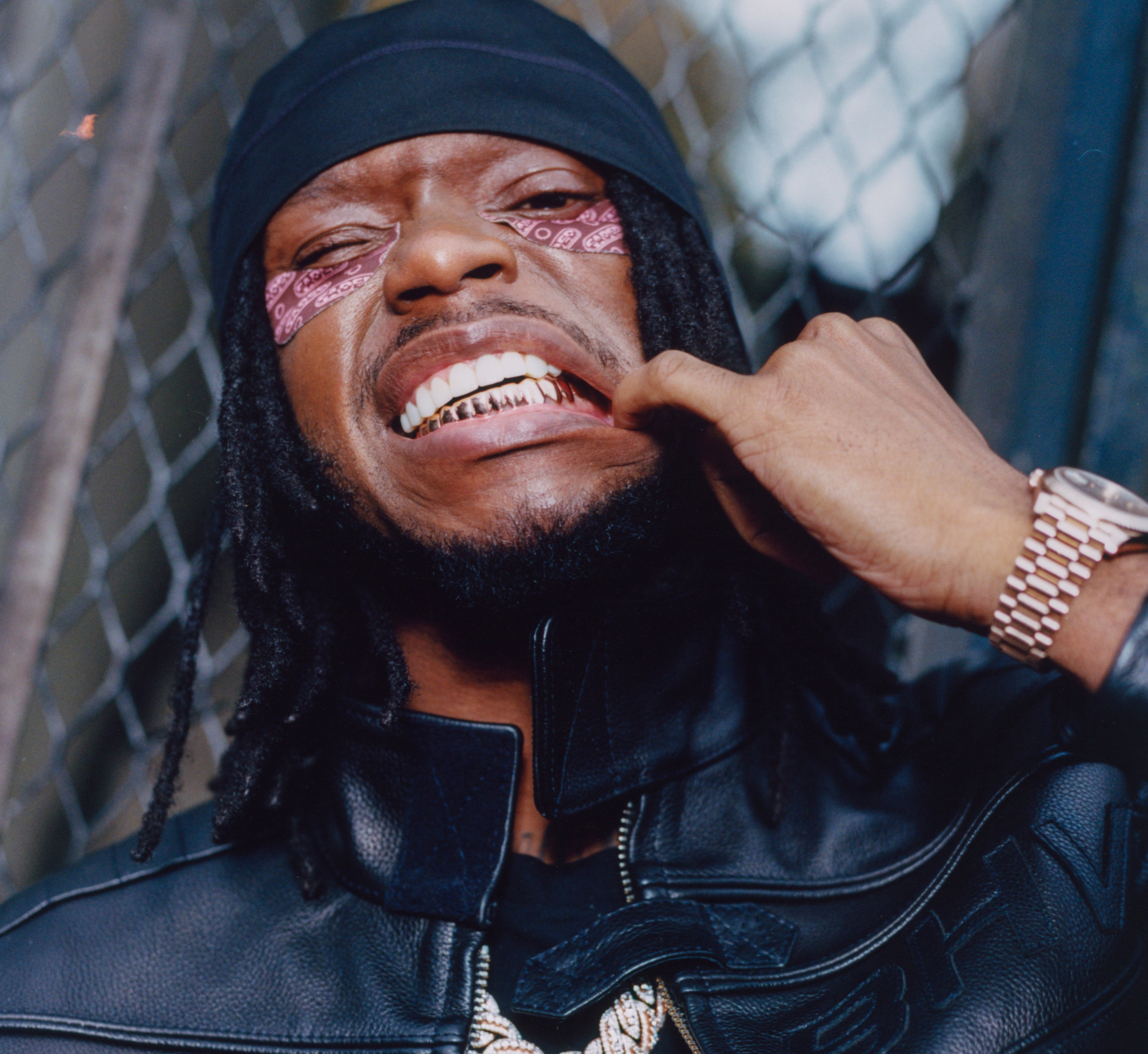 Brightening under eye patches from Topicals Faded in the brown colorway worn by a smiling man with locs, a durag, gold grillz, and a rose gold watch leaning against a chain-link fence.